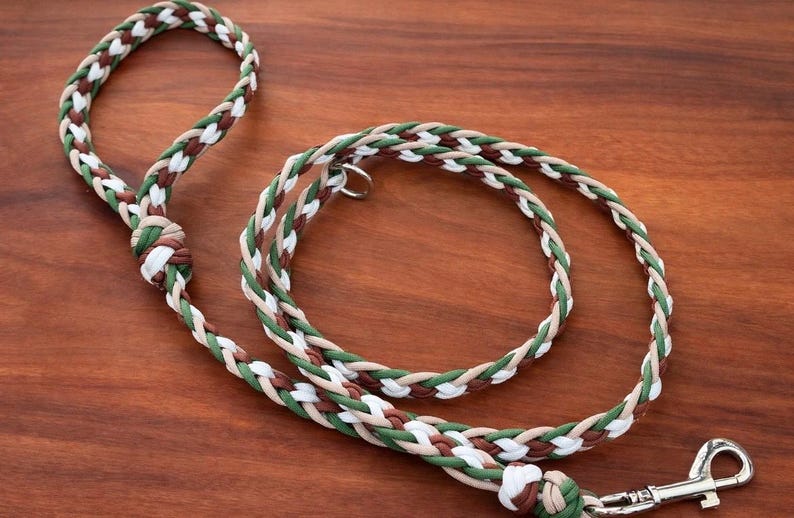 May include: Braided dog leash in shades of green, brown, white, and tan. The leash features a loop handle and a metal clasp. The leash is lying on a wooden surface.