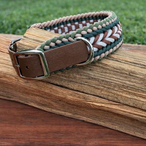 May include: A dog collar with a brown leather strap and a silver-colored buckle. The collar is made of woven cord in shades of green, white, and brown. The collar is resting on a piece of wood.