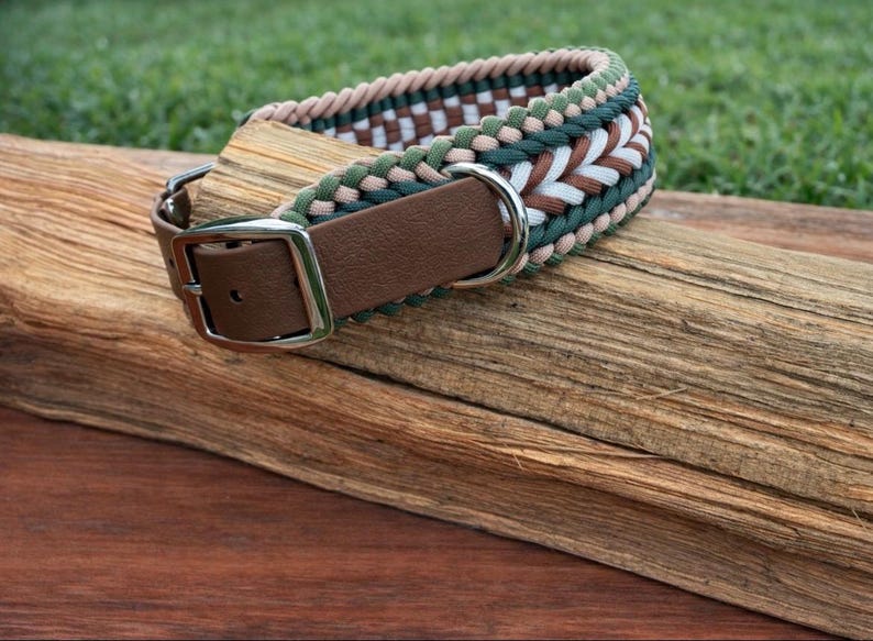 May include: A dog collar with a brown leather strap and a silver-colored buckle. The collar is made of woven cord in shades of green, white, and brown. The collar is resting on a piece of wood.