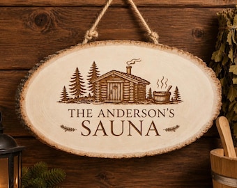 May include: Oval wooden sign with the text "THE ANDERSON'S SAUNA" engraved in a rustic font. The sign features a detailed illustration of a sauna cabin, trees, and a sauna bucket. It hangs from a natural rope.