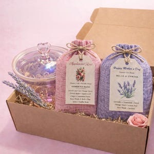 May include: A gift box containing a glass jar with a heart-shaped handle, two fabric pouches, and dried botanicals. The pouches are labeled "Applewood Rose" and "Happy Mother's Day" with ingredient lists. A pink rose is also in the box.