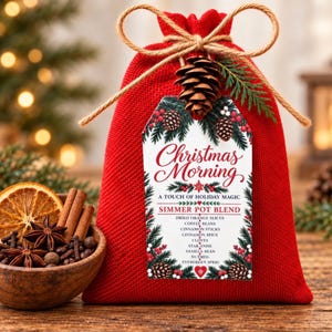 May include: A red fabric drawstring bag tied with twine and a pinecone, featuring a tag labeled Christmas Morning Simmer Pot Blend with a list of ingredients including orange slices, coffee beans, cinnamon, cloves, star anise, and nutmeg.