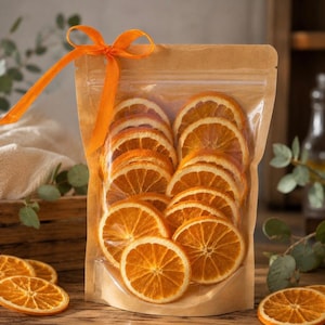 May include: A clear plastic pouch with a brown paper backing containing multiple dehydrated orange slices. The pouch is sealed at the top and features a bright orange ribbon tied in a bow at the upper left corner, showcasing the circular fruit.