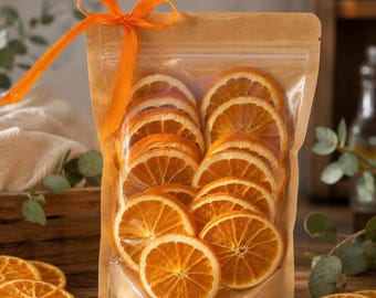 May include: A clear plastic pouch with a brown paper backing containing multiple dehydrated orange slices. The pouch is sealed at the top and features a bright orange ribbon tied in a bow at the upper left corner, showcasing the circular fruit.