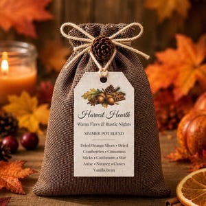 May include: A brown burlap drawstring pouch tied with twine and a small pinecone. A white tag is attached, reading: Harvest Hearth, Warm Fires & Rustic Nights, SIMMER POT BLEND, Dried Orange Slices, Dried Cranberries, Cinnamon Sticks, Cardamom, Star Anise, Nutmeg, Cloves, Vanilla Bean.