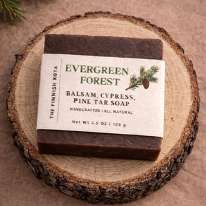 May include: A dark brown bar of soap labeled "Evergreen Forest" with text indicating it contains balsam, cypress, and pine tar. The soap is on a rustic wooden slice. Net weight is 4.5 oz (128 g).