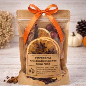 May include: A clear bag filled with dried oranges, cranberries, and spices, tied with an orange ribbon. The label reads "Pumpkin Spice Simmer Pot Kit" with a list of ingredients. The bag is on a light wood surface.