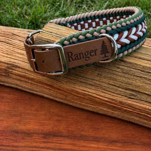May include: A dog collar with a brown leather buckle and a woven design in green, white, brown, and tan. The leather tag is engraved with the word "Ranger" and a tree graphic. The collar is resting on a piece of wood.