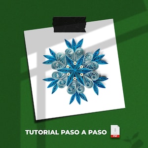 Op de afbeelding: Een handgemaakte papieren sneeuwvlok in blauw- en wittinten, bevestigd op wit papier met zwarte tape. De sneeuwvlok staat centraal, tegen een groene achtergrond. De tekst "TUTORIAL PASO A PASO" is onderaan zichtbaar.