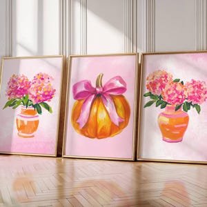 Könnte beinhalten: Drei gerahmte Aquarellmalereien mit goldenen Rahmen. Die Gemälde zeigen einen Kürbis mit einer rosa Schleife und Vasen mit rosa Blumen. Der Hintergrund ist rosa. Die Gemälde lehnen an einer weißen Wand.