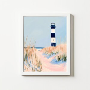 Puede incluir: Una impresión artística enmarcada que presenta una escena costera. Un faro a rayas blancas y negras se alza sobre una duna de arena, con hierbas altas y un camino que conduce al océano bajo un cielo azul claro. La obra de arte está en un marco blanco.