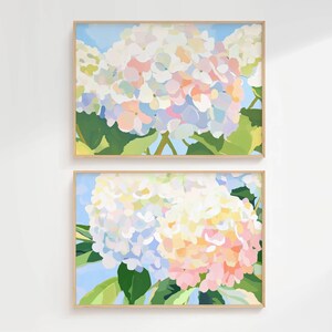 K&ouml;nnte beinhalten: Zwei gerahmte florale Kunstdrucke. Jeder Druck zeigt eine Nahaufnahme einer Hortensienbl&uuml;te in Wei&szlig;-, Rosa- und Blaut&ouml;nen vor einem hellblauen Hintergrund. Das Kunstwerk ist in hellem Holz gerahmt.