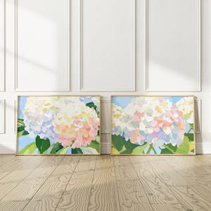 K&ouml;nnte beinhalten: Zwei gerahmte Kunstdrucke mit Blumenmotiven. Jeder Druck zeigt einen farbenfrohen Hortensienstrau&szlig; in Rosa-, Gelb- und Blaut&ouml;nen vor einem hellblauen Hintergrund. Die Rahmen sind hellbraun und das Kunstwerk wird auf einem Holzboden pr&auml;sentiert.