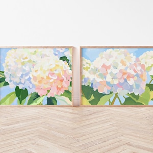May include: Two framed floral art prints. Each features a close-up of a hydrangea bloom in shades of white, pink, and blue against a light blue background. The artwork is framed in a light wood frame, creating a cohesive set.