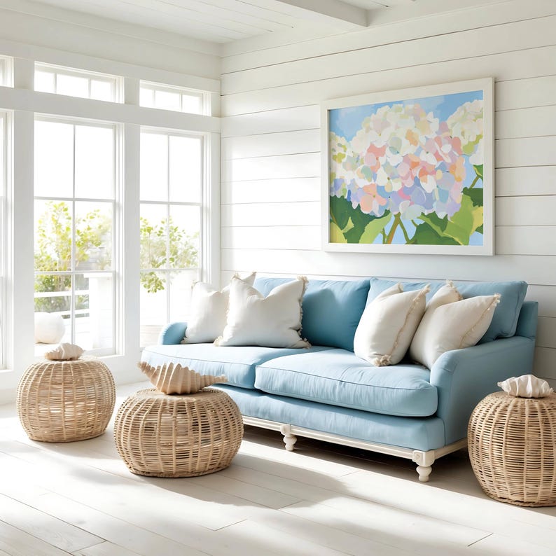K&ouml;nnte beinhalten: Ein hellblaues Sofa mit wei&szlig;en Kissen und einem gerahmten Blumenbild. Rattan-Hocker mit dekorativen Muscheln stehen vor dem Sofa. Der Raum hat wei&szlig;e W&auml;nde und gro&szlig;e Fenster, die eine helle und luftige Atmosph&auml;re schaffen.