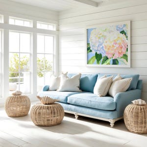 K&ouml;nnte beinhalten: Hellblaues Sofa mit wei&szlig;en Kissen und einem gerahmten Blumenbild. Rattan-Hocker und dekorative Muscheln erg&auml;nzen das K&uuml;sten-Thema. Der Raum hat wei&szlig;e W&auml;nde und gro&szlig;e Fenster.
