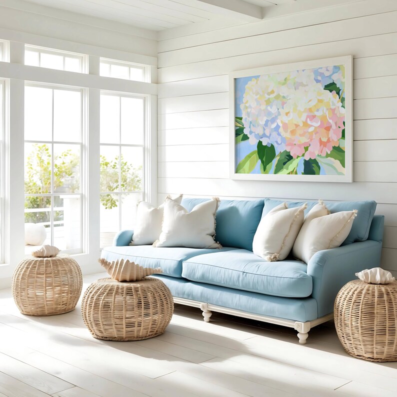 K&ouml;nnte beinhalten: Hellblaues Sofa mit wei&szlig;en Kissen und einem gerahmten Blumenbild. Rattan-Hocker und dekorative Muscheln erg&auml;nzen das K&uuml;sten-Thema. Der Raum hat wei&szlig;e W&auml;nde und gro&szlig;e Fenster.