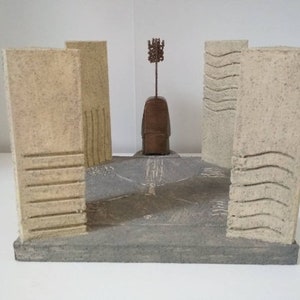 Pode incluir: Um modelo em miniatura de uma estrutura de pedra com quatro paredes e uma figura central. As paredes são texturizadas e têm uma cor bege claro. A figura central é de forma redonda e marrom com uma ponta de metal no topo.