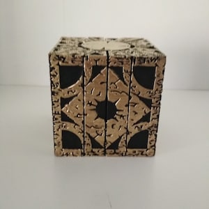 Puede incluir: Un cubo negro con detalles dorados y patrones intrincados. El cubo tiene una superficie texturizada y parece estar hecho de metal.