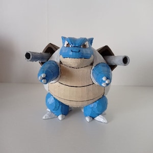 Könnte beinhalten: Eine blaue und braune 3D-gedruckte Figur eines Pokemon-Charakters, Blastoise. Die Figur hat eine blaue Schale mit braunen Akzenten und einen weißen Unterbauch. Sie hat zwei Kanonen auf dem Rücken.