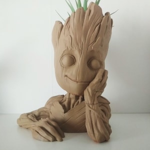 Puede incluir: Un macetero de cerámica marrón con forma del personaje Groot de la película Guardianes de la Galaxia. El macetero tiene una pequeña planta verde que crece en la parte superior de su cabeza.