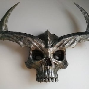 Puede incluir: Una máscara de calavera plateada y gris con cuernos grandes y curvos. La máscara tiene rasgos faciales detallados, incluyendo cuencas de los ojos, una nariz y dientes. La máscara es probablemente para disfraces o fines decorativos.