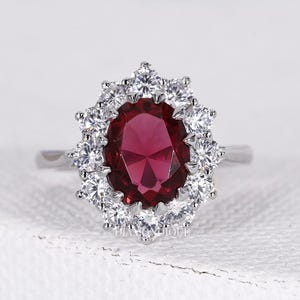 Könnte beinhalten: Ein silberner Ring mit einem großen, oval geschliffenen, tiefroten Edelstein, umgeben von einem Kranz aus kleineren, klaren Steinen. Das Design des Rings ist klassisch und elegant, wobei der Fokus auf der satten Farbe und dem Glanz des zentralen Edelsteins liegt.