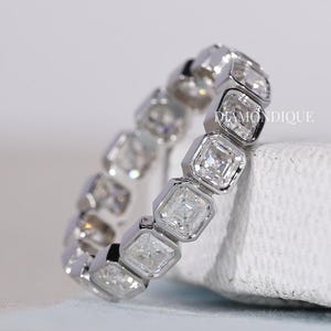 Könnte beinhalten: Ein silberner Eternity-Ring mit quadratisch geschliffenen, klaren Edelsteinen. Der Ring ist vor einem weichen, weißen Hintergrund platziert, mit dem Markennamen "DIAMONDIQUE" sichtbar. Der Ring ist ein Schmuckstück.