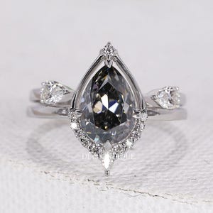 Könnte beinhalten: Ein silbernes Verlobungsring-Set mit einem birnenförmigen, grau getönten Edelstein als Mittelpunkt. Der Ring ist mit kleineren, klaren Edelsteinen und einem zarten Band verziert. Das Set enthält ein passendes Band mit zusätzlichen kleinen Edelsteinen.