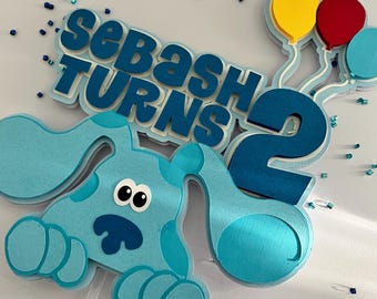 Puede incluir: Un adorno para pastel con temática de Blue's Clues que presenta un perro azul con ojos grandes y una nariz azul. El adorno también incluye las palabras "Sebash Turns 2" y tres globos en rojo, amarillo y azul.