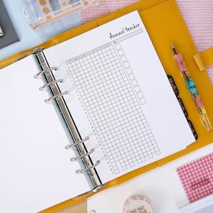 Puede incluir: Una carpeta amarilla con una página blanca abierta a un rastreador anual imprimible. El rastreador tiene una cuadrícula con columnas para cada mes del año y filas para cada día del mes. El texto "Annual Tracker" está escrito en la parte superior de la página.