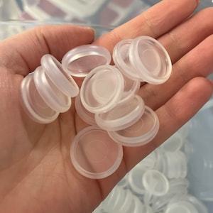 May include: A hand holding a pile of clear plastic round discs. The discs are about 1 inch in diameter and have a slightly raised edge.