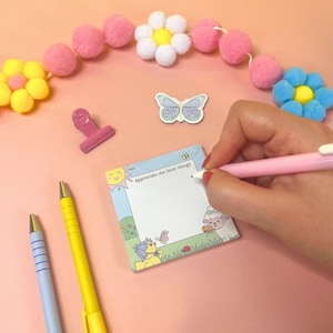 May include: A pink pen writing on a small notepad with the text "Appreciate the little things." The notepad features a cartoon illustration of a sheep, butterfly, and sun. A pink paper clip, butterfly sticker, and pens are also visible.