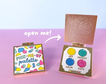 Puede incluir: Un pin "Pick-Me-Up Palette" con marco dorado y tapa rosa dorado brillante. El pin cerrado presenta formas abstractas coloridas y texto. El pin abierto revela cuatro círculos brillantes con las palabras "Joy", "Energy", "Calm" y "Hope".