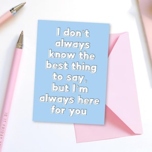 Op de afbeelding: Een lichtblauwe wenskaart met de tekst "I don't always know the best thing to say, but I'm always here for you." De kaart ligt naast een roze envelop en twee roze pennen.