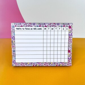 Puede incluir: Un rastreador de hábitos rectangular con un borde floral. El rastreador tiene un interior blanco con líneas para rastrear los hábitos y los días de la semana. El texto "Habits to focus on this week" está en la parte superior. El borde es una mezcla de diseños florales rosas, morados y verdes.