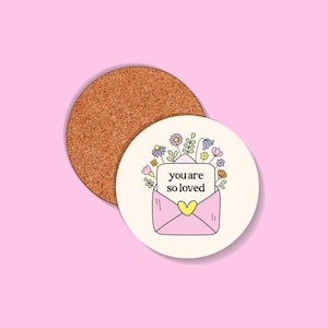 Può includere: Un sottobicchiere rotondo in sughero accanto a un sottobicchiere bianco con un disegno di busta rosa. La busta ha un cuore giallo e le parole "you are so loved". Il design include fiori colorati.