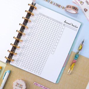 Pu&ograve; includere: Un planner annuale bianco con una griglia per ogni mese dell'anno. Il planner ha una sezione per gli appunti e un titolo che dice "Annual Tracker".