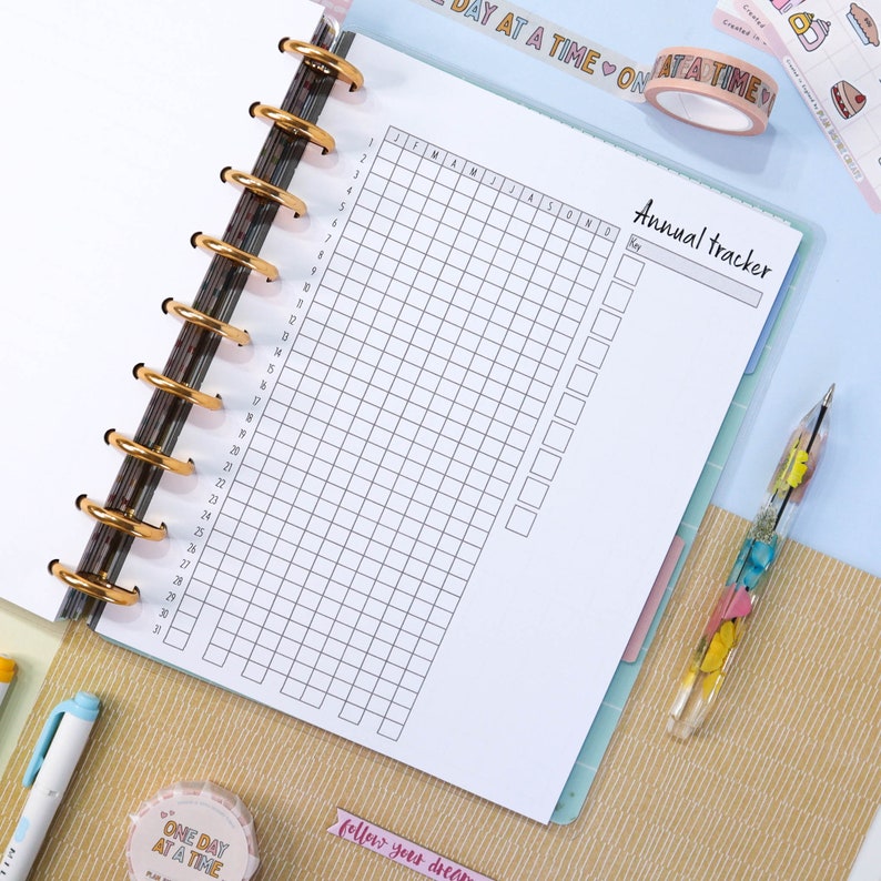 Pu&ograve; includere: Un planner annuale bianco con una griglia per ogni mese dell'anno. Il planner ha una sezione per gli appunti e un titolo che dice "Annual Tracker".