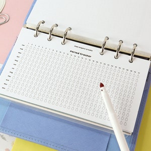 May include: A white period tracker page with a grid of raindrops for each day of the year. The page is in a blue binder with silver rings. A white pen with a red tip is on the page.