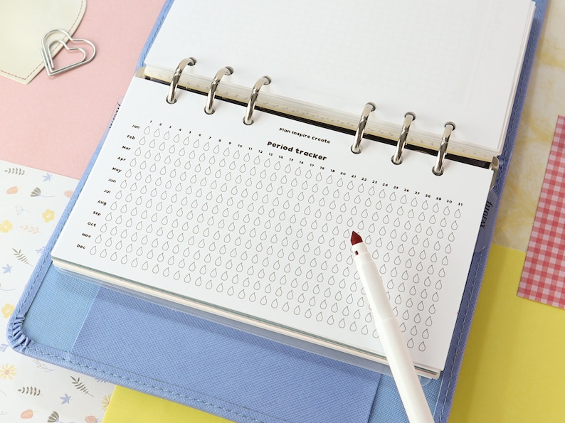 May include: A white period tracker page with a grid of raindrops for each day of the year. The page is in a blue binder with silver rings. A white pen with a red tip is on the page.