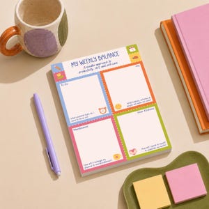 May include: A rectangular paper notepad titled My Weekly Balance with sections for To Do, Joy, Maintenance, and Inner Kindness. The page features colorful borders, small icons, and prompts for productivity and self-care. A light purple pen lies nearby.