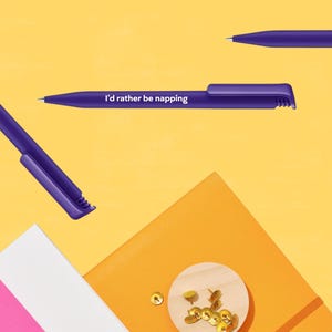 Puede incluir: Tres bolígrafos morados sobre un fondo amarillo. Un bolígrafo tiene el texto blanco "I'd rather be napping" impreso en el cuerpo. Los bolígrafos tienen un clip y una punta cónica. Debajo se observa un pequeño disco de madera con chinchetas doradas.