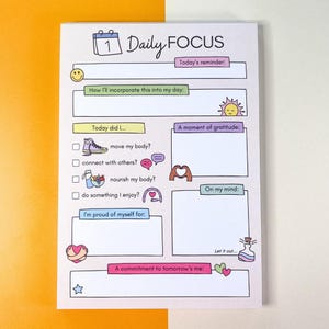 以下が含まれることがあります： 淡いピンク色の背景とカラフルなイラストが特徴の「Daily Focus」ノートパッド。リマインダー、毎日の目標、感謝、コミットメントのセクションが含まれています。「Daily Focus」、「Today's reminder」、「A commitment to tomorrow's me」などのテキストが含まれています。