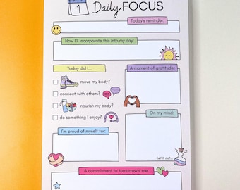 May include: A Daily Focus notepad with a light pink background and colorful illustrations. The notepad includes sections for reminders, daily goals, gratitude, and commitments. The text includes "Daily Focus", "Today's reminder", and "A commitment to tomorrow's me".