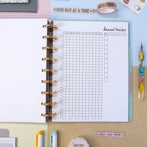Pu&ograve; includere: Una pagina bianca con un calendario a griglia per tenere traccia degli obiettivi durante tutto l'anno. Il calendario &egrave; etichettato "Annual Tracker" e include i mesi dell'anno, nonch&eacute; i giorni numerati. La pagina fa parte di un quaderno rilegato con anelli dorati.