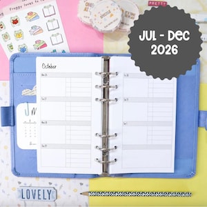 Op de afbeelding: Blauwe planner met een wekelijkse indeling, met de tekst "October" en data. Inclusief stickers, decoratieve tape rollen en een potlood. De planner toont "JUL - DEC 2026" op een grijs cirkelvormig label.