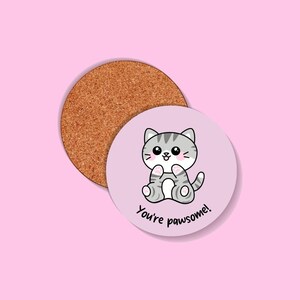 Può includere: Due sottobicchieri rotondi. Uno è in sughero, l'altro è lilla chiaro con un gatto tabby grigio dei cartoni animati e la scritta "You're pawsome!". Il gatto ha un'espressione felice ed è seduto.