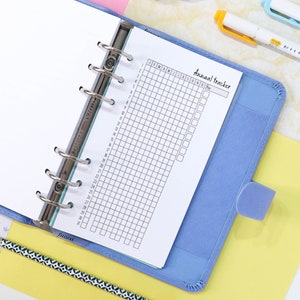 Pu&ograve; includere: Un raccoglitore blu con una pagina bianca aperta su un tracker mensile. La pagina &egrave; etichettata "Tracker annuale" e ha una griglia con caselle per ogni mese dell'anno. La pagina ha anche una sezione per una chiave.