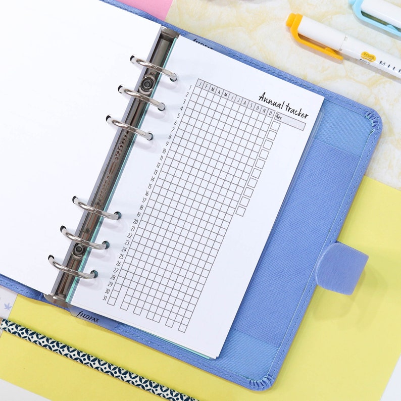 Pu&ograve; includere: Un raccoglitore blu con una pagina bianca aperta su un tracker mensile. La pagina &egrave; etichettata "Tracker annuale" e ha una griglia con caselle per ogni mese dell'anno. La pagina ha anche una sezione per una chiave.